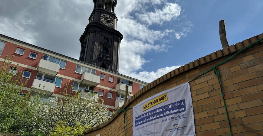 Ein Banner hängt an der Mauer einer Kita. Im Hintergrund ist der Turm der St. Michaelis Kirche in Hamburg zu sehen - Copyright: Christian Schierwagen