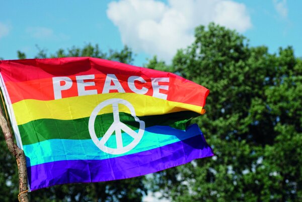 Flagge in Querstreifen (Regenbogenfarben) mit Peace-Zeichen vor grünen Bäumen und blauem Himmel - Copyright: Ben Frieden, Pixabay